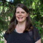 Head shot portrait of Kara Haas wearing a dark shirt against a background of conifers.