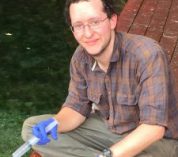 Robert Logan sits on a bench wearing blue latex gloves and holding a syringe.