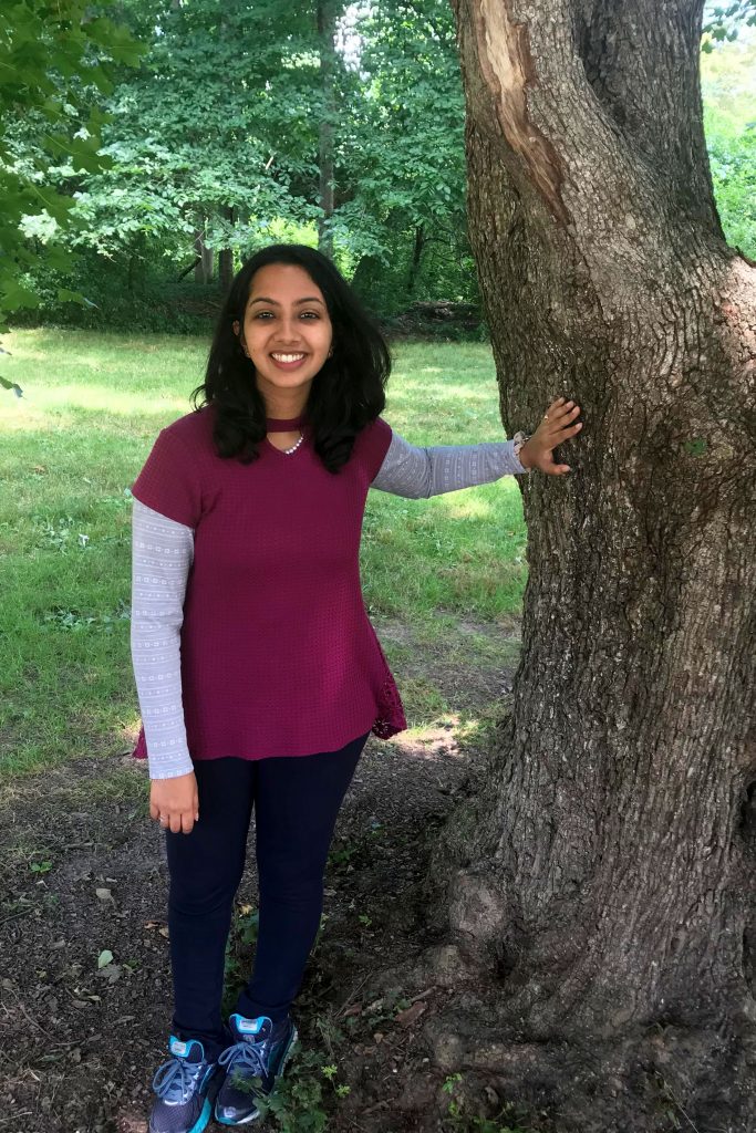 Akshata Rudrapatna stands with her hand resting against a tree trunk at the Kellogg Biological Station.