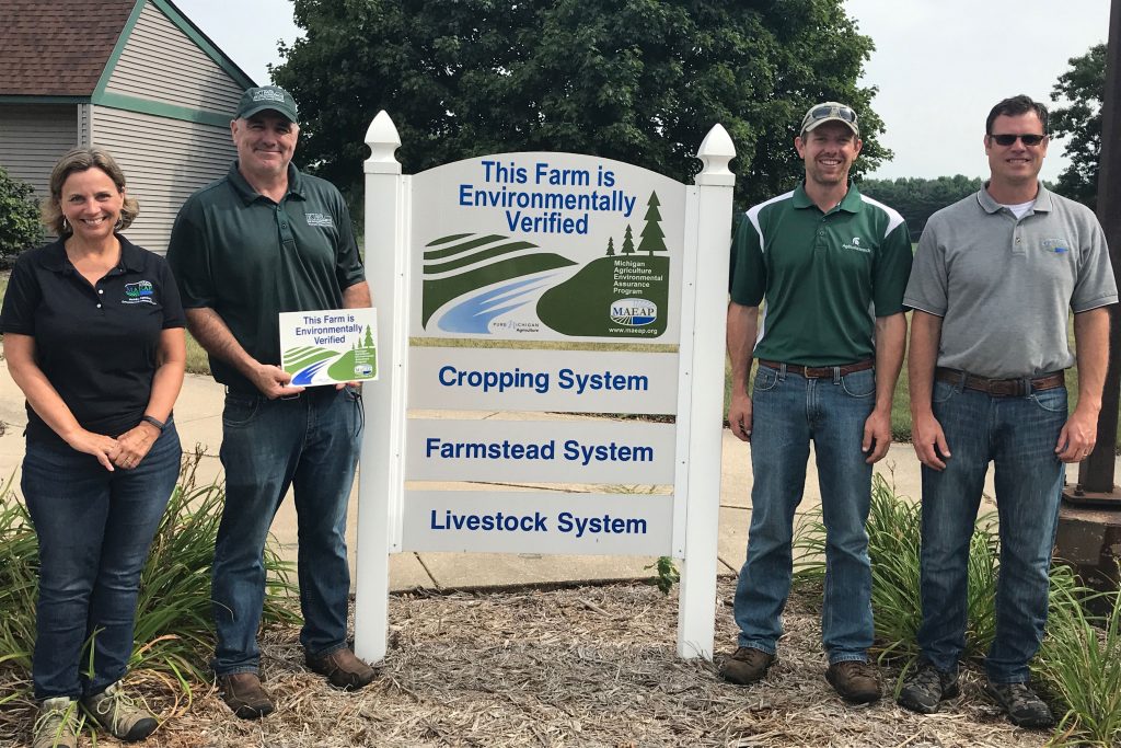 Two State of Michigan agriculture and rural development staff members stand with Kellogg Farm managers Straub and Wilke around the Kellogg Farm's environmental verification sign.
