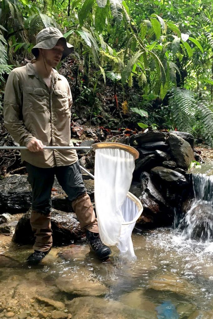 Kyle Jaynes takes notes holds a fish net while standing in a stream in Trinidad.