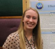 Sydney Hall stands, smiling, next to her research poster at the KBS Undergraduate Research Symposium.