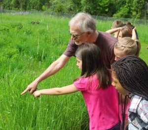 A KBS volunteers points at something on the ground in a grassy area while a group of schoolchildren look on.