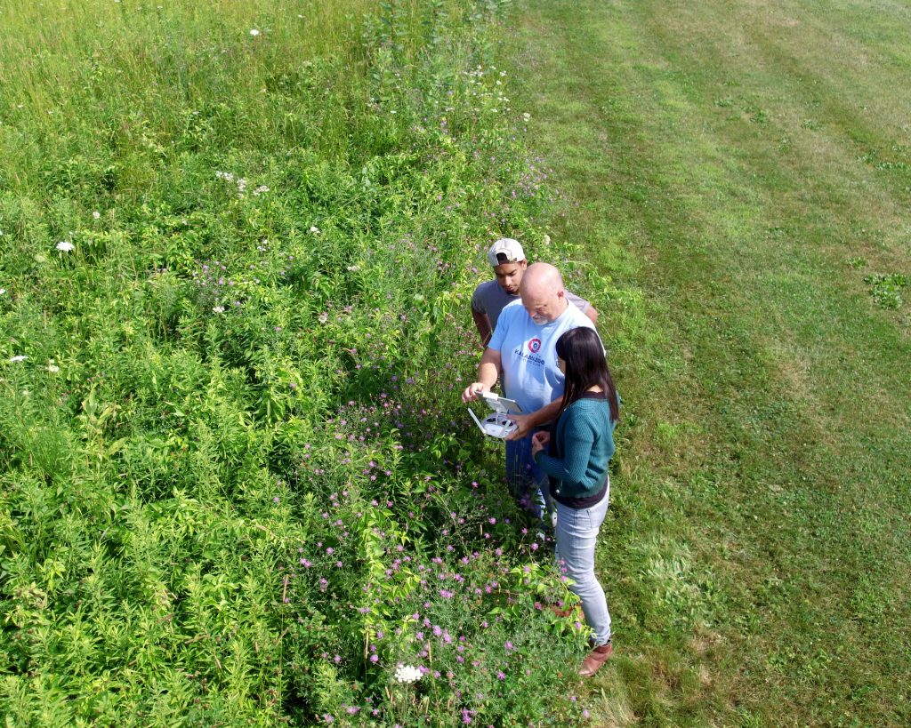 Brandon D'Souza, Kevin Kahmark, and Dr. Hsunyi Hsieh examine a drone while standing at the edge of a research field.
