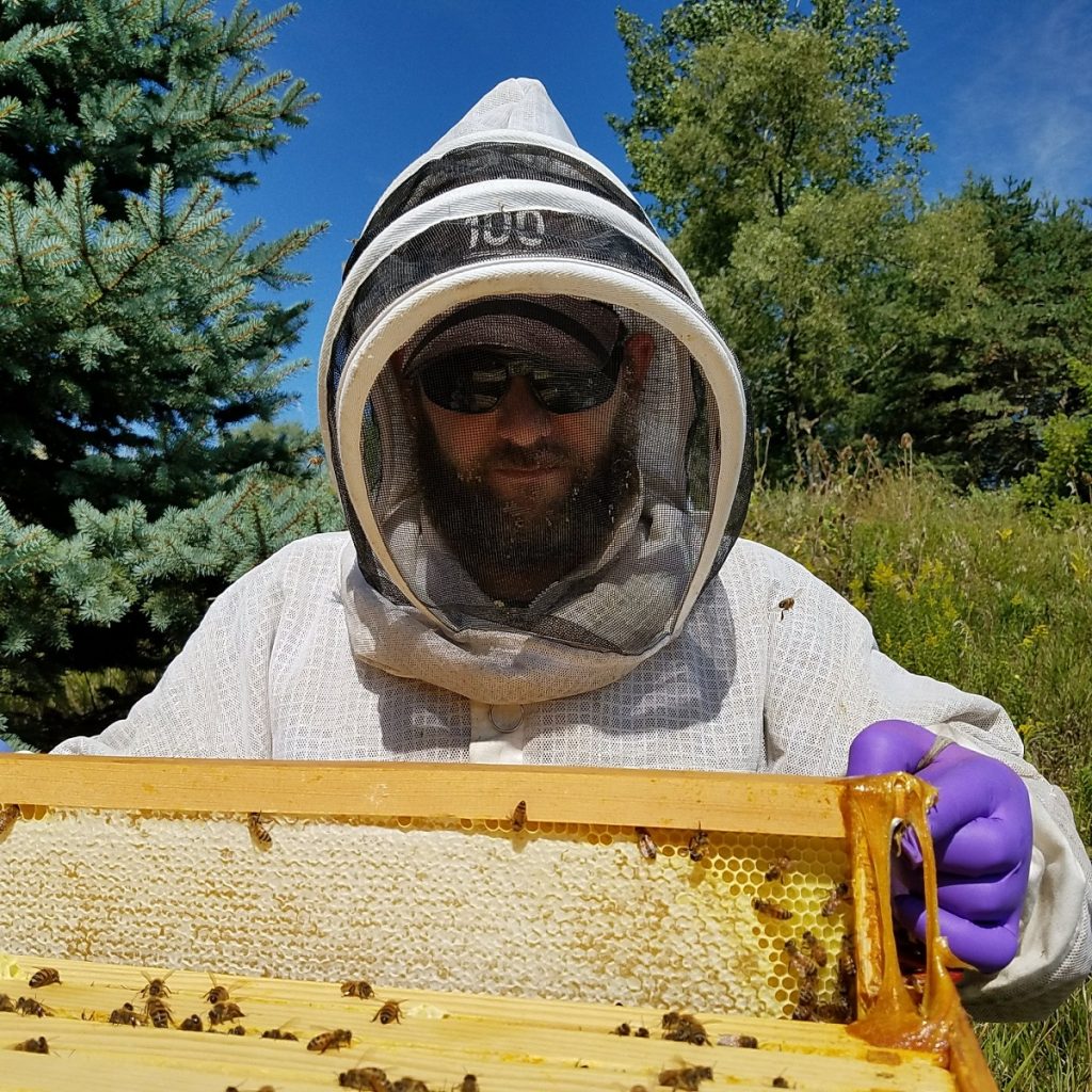 Adam Ingrao, wearing a white beekeeping suit, holds a frame from a honey bee hive.