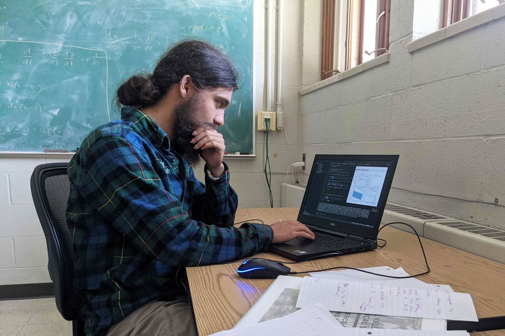Joseph Savage sits in a classroom, focused on working on his computer. Equations are written on a chalkboard in the background.