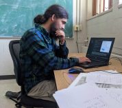 Joseph Savage sits in a classroom, focused on working on his computer. Equations are written on a chalkboard in the background.