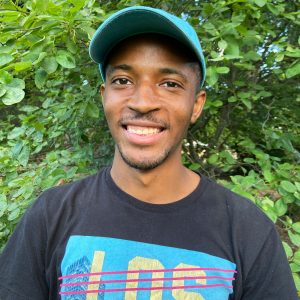 Ceco Maples, wearing a black T-shirt and a blue baseball cap, smiles at the camera against a background of trees and shrubs.