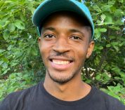 Ceco Maples, wearing a black T-shirt and a blue baseball cap, smiles at the camera against a background of trees and shrubs.