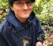 Glade Dlott stands in a wooded area wearing a jacket and hat, looking at the camera and holding two dark-colored mushrooms.