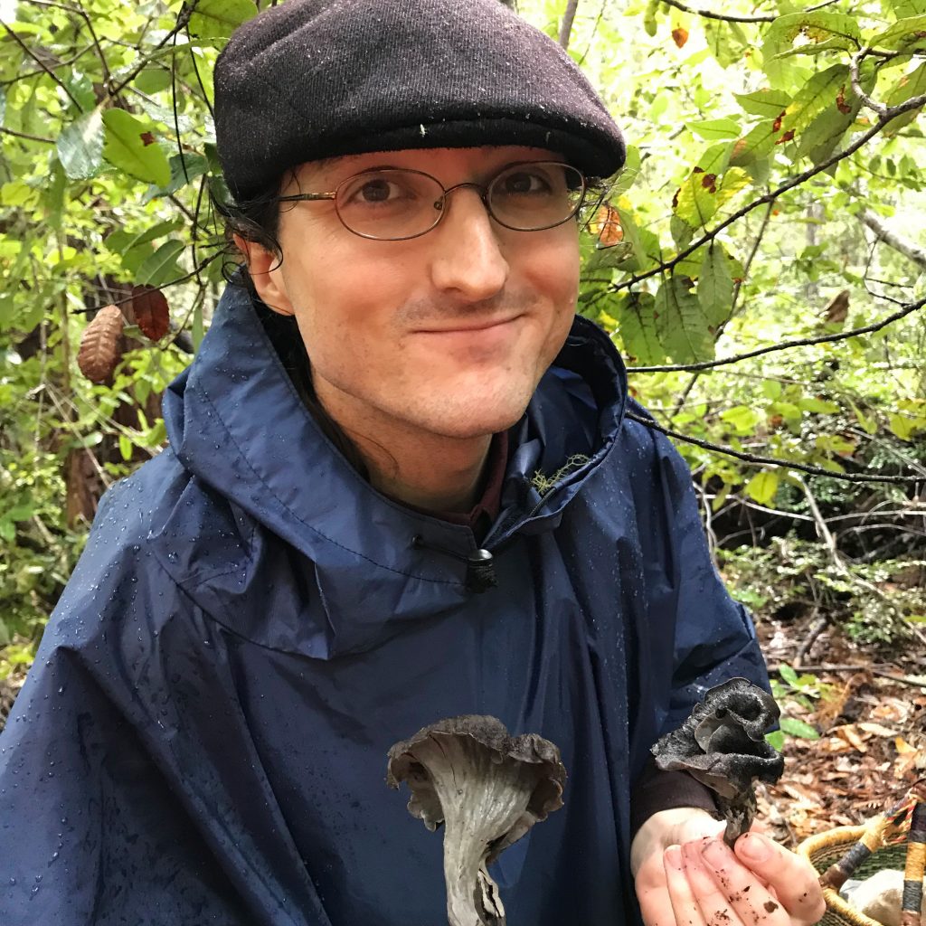 Glade Dlott stands in a wooded area wearing a jacket and hat, looking at the camera and holding two dark-colored mushrooms.
