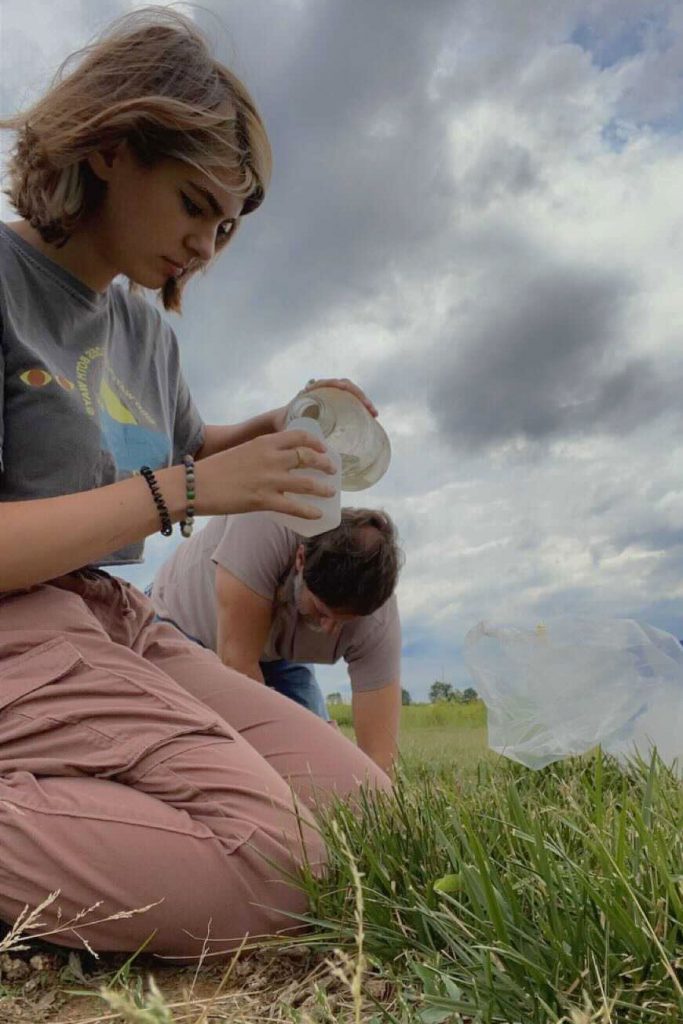 Against a cloudy sky, Katherine Egeler pours a bromide solution from one bottle to another, in preparation for watering switchgrass plots.