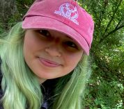 Cheyenne Cope, wearing a pink baseball cap, smiles at the camera while standing in a wooded area.