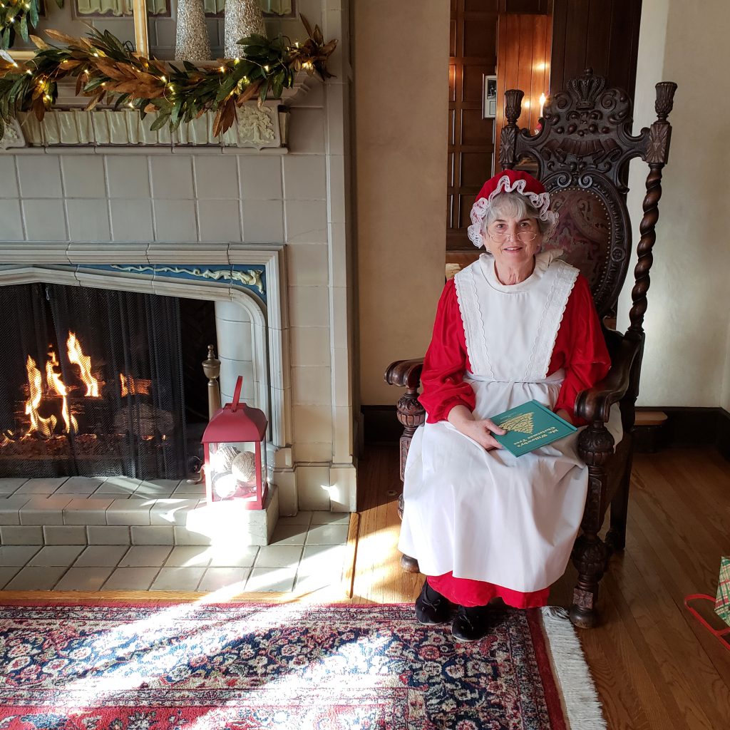 Mrs. Claus sits in an ornate chair beside a lit fireplace in the W.K. Kellogg Manor House, holding a book on her lap.
