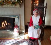 Mrs. Claus sits in an ornate chair beside a lit fireplace in the W.K. Kellogg Manor House, holding a book on her lap.