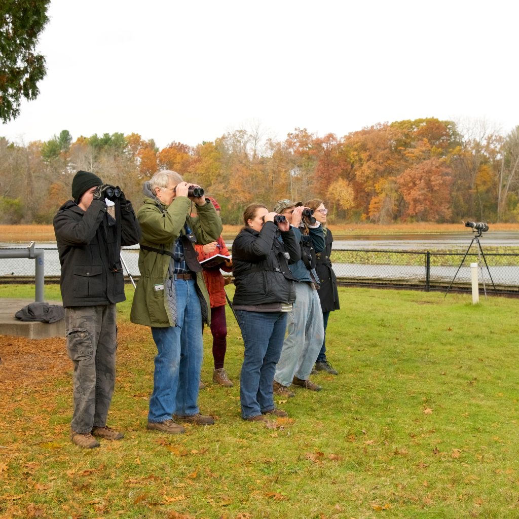 A group of people stand together, peering through binoculars, on the shores of Wintergreen Lake with trees in autumn color in the background.