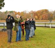 A group of people stand together, peering through binoculars, on the shores of Wintergreen Lake with trees in autumn color in the background.