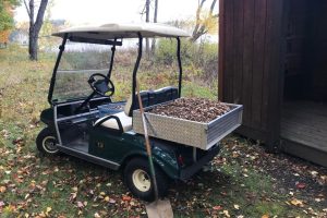 A green golf cart donated to the Kellogg Bird Sanctuary, with the bed filled with wood chips, is parked near the Sanctuary's bird blind, with Wintergreen Lake visible in the background.