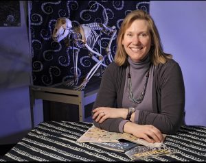 Head shot of Anne Bronikowski seated at a table with a stack of magazines.