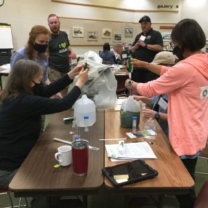 Jake Nalley, wearing an "I love algae" T-shirt, looks on as four people seated at a table use droppers to add liquid to milk jugs.