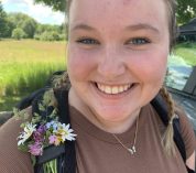 Alex Sills smiles at the camera, wearing a backpack with picked flowers tucked into the shoulder strap.