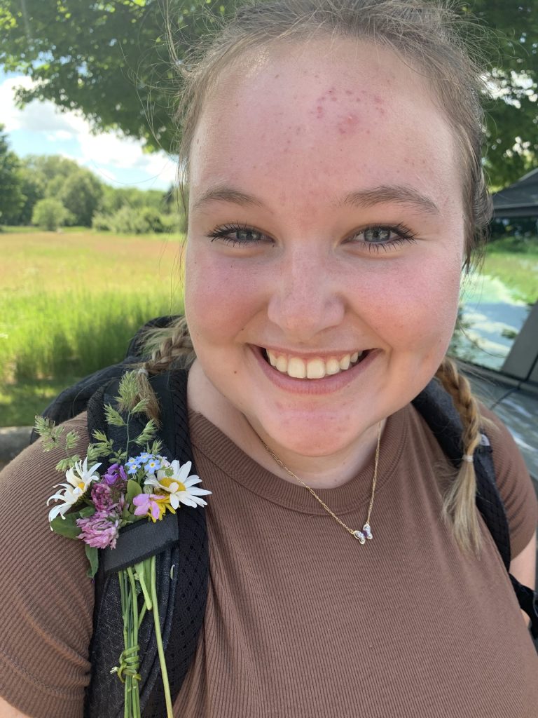 Alex Sills smiles at the camera, wearing a backpack with picked flowers tucked into the shoulder strap.