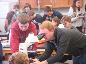 Jake Nalley peers into a microscope in a lab, surrounded by high school students.