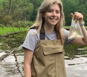 Marie Kroneman stands in the water holding a water sample in a bag.