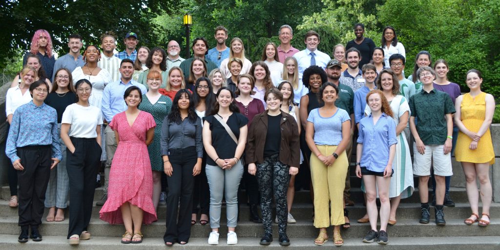 W.K. Kellogg Biological Station 2022 summer students and mentors pose for a group photo on a set of stairs with trees in the background.
