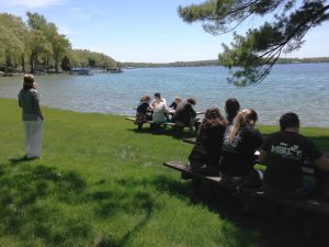 Educators sit at two picnic tables on a green lawn on the shore of Gull Lake.