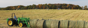 A green and yellow tractor pushes large round bales of greenish brown switchgrass on a flat, golden field. In the distance, trees begin to show the auburn colors of autumn under a blue sky streaked with large white clouds.