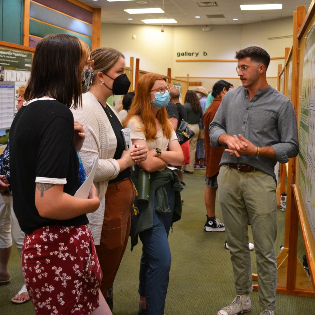 An undergraduate student stands with folded hands while answering questions for three symposium attendees.