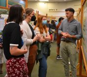 An undergraduate student stands with folded hands while answering questions for three symposium attendees.
