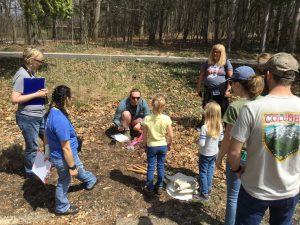 A group of adults and children gather around a person crouching with a clipboard, during a survey of insects.