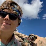 Selfie photo of Caitlin Broderick, standing in front of a rock formation with a blue sky and canine companion in the background.