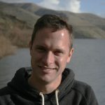 Christopher P. Kozakiewicz stands in front of a body of water and a mountainous background, on a sunny day.