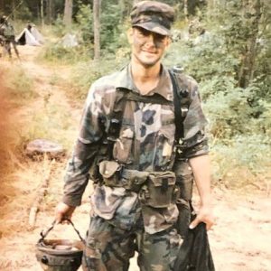 Jim Allen stands on a trail in a forested area, wearing fatigues and holding his helmet, during a deployment to Iraq.