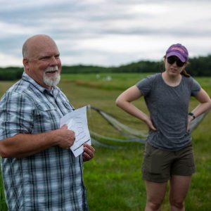 Kevin Kahmark points to a diagram on a sheet of paper with another person looking on, while standing in a field at the W.K. Kellogg Biological Station.
