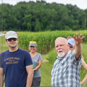 Kevin Kahmark gestures at some equipment in the foreground while a small group of people look on, while standing in a field at the W.K. Kellogg Biological Station.