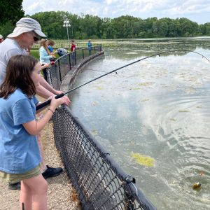 Several people stand along the fence bordering Wintergreen Lake at the W.K. Kellogg Bird Sanctuary, fishing.
