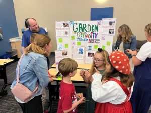 Students examine bracelets they made while standing in front of a poster about a class project on garden diversity.