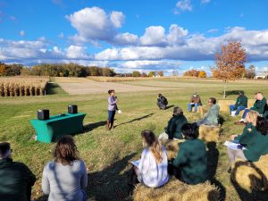 Alisha Shah speaks to the group from the inaugural Spartan Bus Tour, who are sitting on straw bales in the foreground, at the KBS Long-term Agroecosystem Research site.