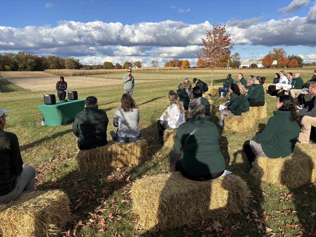 MSU President Kevin Guskiewicz speaks to the group from the inaugural Spartan Bus Tour, who are sitting on straw bales in the foreground, at the KBS Long-term Agroecosystem Research site.