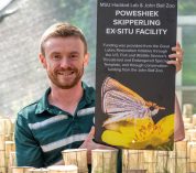 David Pavlik poses in a greenhouse at John Ball Zoo with an informational placard on his research project with Poweshiek skipperlings.