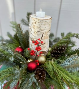 A holiday arrangement featuring evergreen boughs, pine cones and tree ornaments with a birch bark candle in the center.