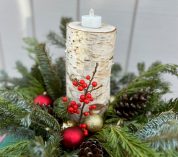 A holiday arrangement featuring evergreen boughs, pine cones and tree ornaments with a birch bark candle in the center.