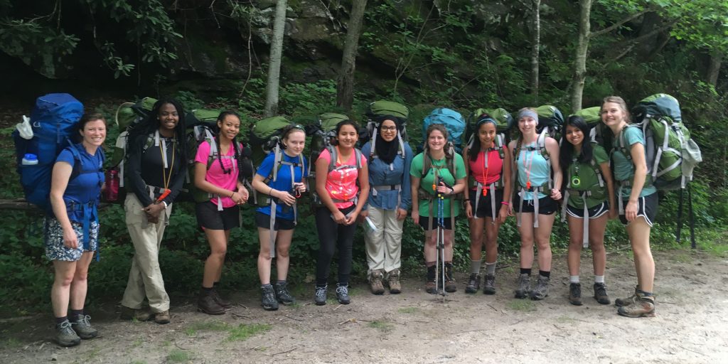 A group of participants in a past GALS program pose with their backpacks for a photo with a forest in the background.