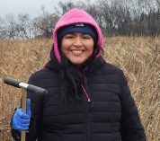 Natali Gonzalez stands in a field in late autumn, holding a tool and wearing a heavy coat and hat.