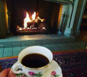 A person holds a cup of tea in an ornate teacup in front of the fireplace in the Manor House living room.