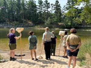 Several GALS participants stand on sand at the water's edge, holding fish nets, while an employee from the Michigan Department of Natural Resources talk with them about aquatic ecology.